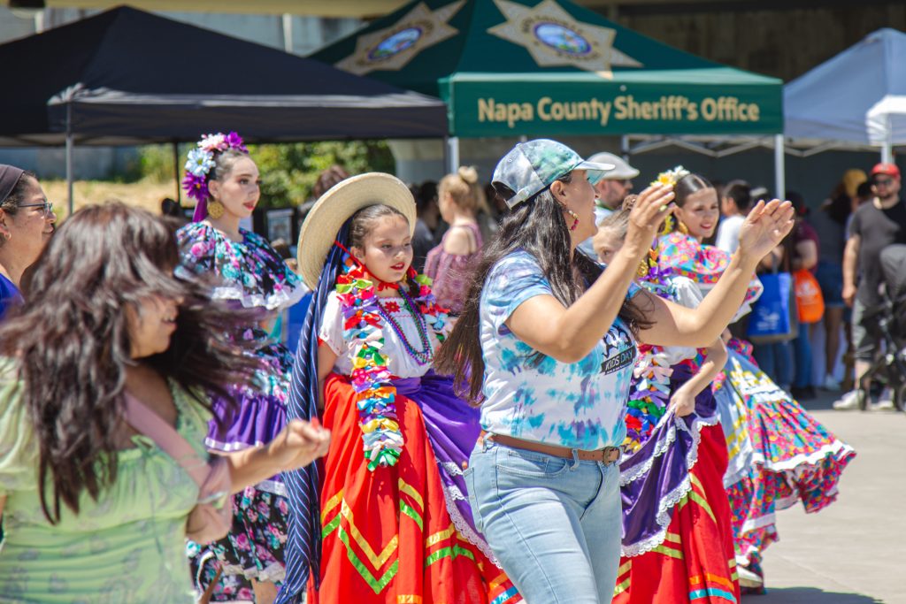 Group of dancers in bright, traditional dresses performing at an outdoor festival, with a Napa County Sheriff's Office tent in the background.
