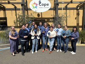 Group of adults posing outside a yellow building under a Cope Family Center sign, holding blue pinwheels and smiling.